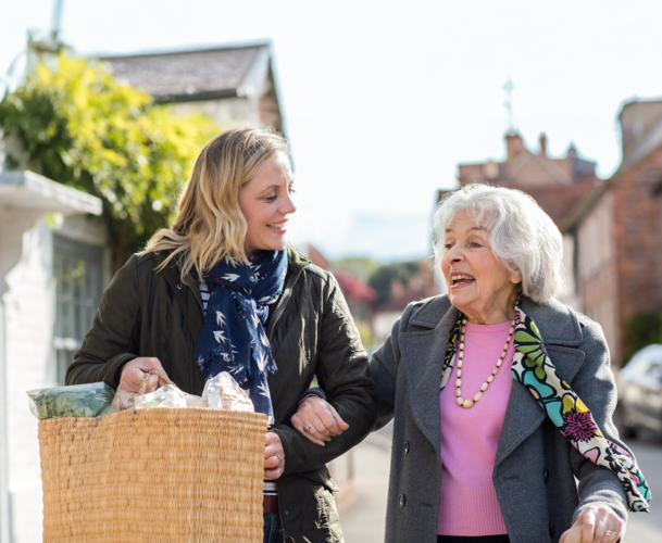 Eine j�ngere Frau tr�gt die Eink�ufe von einer �lteren Frau und begleitet sie.